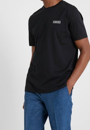 Man draagt een zwart T-shirt met een klein wit omgekeerd "HUGO" logo op de borst en een blauwe broek, zijn rechterhand in de zak.