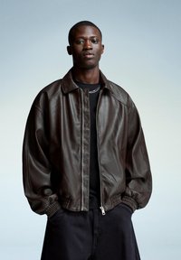 Brown leather bomber jacket with a zip front, ribbed cuffs, and a relaxed fit over a black shirt. Background features a soft gradient.