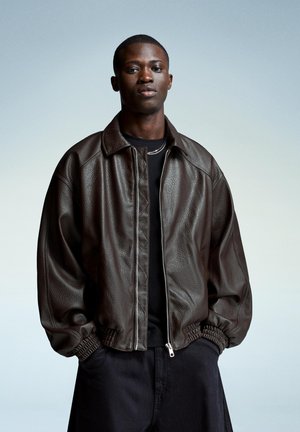 Brown leather bomber jacket with a zip front, ribbed cuffs, and a relaxed fit over a black shirt. Background features a soft gradient.