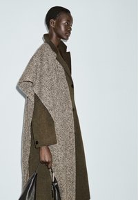Cappotto oversize verde oliva e beige con una finitura texture, caratterizzato da grandi bottoni e un design stratificato unico, accompagnato da una borsa nera.