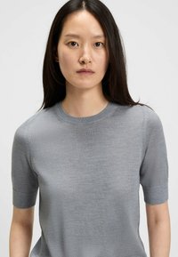 Maglietta a maniche corte di colore grigio chiaro, realizzata in un tessuto morbido, con scollatura tonda e una texture liscia con una sottile lucentezza.