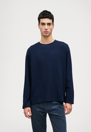 Navy blue long-sleeve crew neck sweater in a textured knit, paired with dark pinstriped trousers. Classic, relaxed fit.