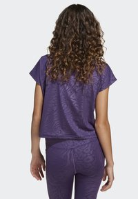 Woman with curly brown hair wearing a purple short-sleeve top and matching leggings with a subtle animal print, facing away.