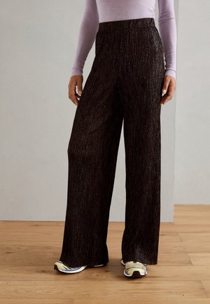 Vrouw draagt wijde, glanzende zwarte broek met beige sneakers en een lichtpaarse lange mouwen top, staand op een houten vloer.