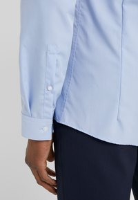Chemise à manches longues bleu clair avec des poignets boutonnés, coupe ajustée et tissu lisse. Présentée avec un pantalon sombre, mettant en avant le contraste des couleurs.