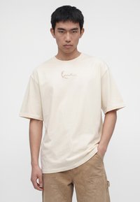 Junger Mann trägt ein lockeres cremefarbenes T-Shirt mit kleinem Logo und hellbraune Cargohosen, steht vor einem schlichten weißen Hintergrund.