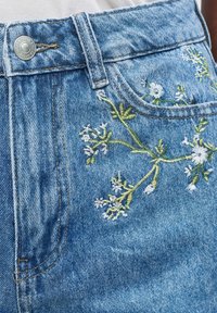 Jean en denim bleu avec broderie florale verte et blanche près de la poche avant et un bouton argenté à la taille.