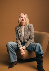 Blazer a motivo scozzese, T-shirt color crema, jeans azzurri chiari, stivali marroni al ginocchio. Modello seduto su una sedia color cuoio con sfondo marrone.