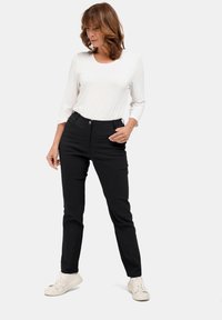 Zwarte, strakke broek met een gladde textuur en voorzakken, gecombineerd met een witte, lange mouw shirt. Model staat met één hand op de heup.
