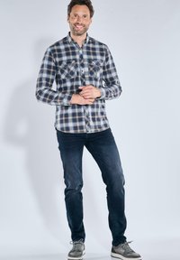 Langärmliges karriertes Hemd in Blau, Marineblau und Weiß; mit zwei Brusttaschen, knöpfbarem Frontverschluss und abgerundetem Saum; kombiniert mit dunklen Jeans und Sneakers.