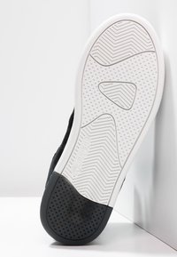 Baskets noires avec une tige texturée, semelle en caoutchouc blanc avec un motif en chevrons, et un logo de marque prominent sur le côté.