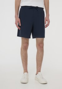 Pantaloni corti blu navy realizzati in tessuto leggero, con un elastico in vita e un cordino, oltre a tasche laterali. Indossati con scarpe da ginnastica bianche.