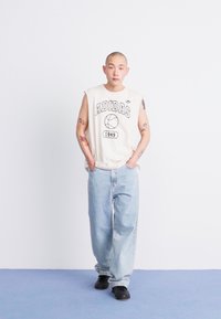 Beige, ujj nélküli Adidas póló fekete felirattal és kosárlabdát ábrázoló grafikával, laza világoskék farmerral és fekete cipővel párosítva. Egyszínű háttér.