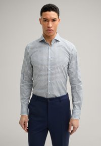 Camicia a maniche lunghe di un blu chiaro con un motivo, realizzata in un tessuto liscio. Indossata con un paio di pantaloni blu navy, dotati di chiusura a bottoni.