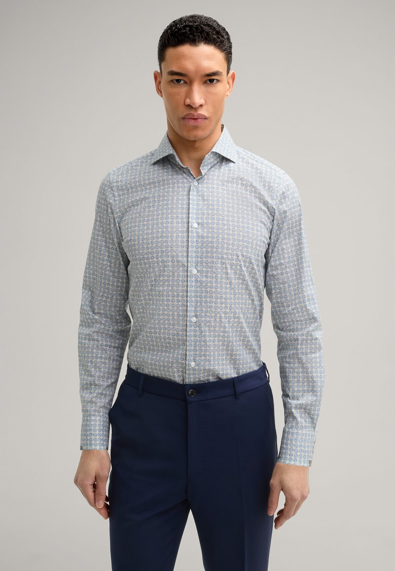 Camicia a maniche lunghe di un blu chiaro con un motivo, realizzata in un tessuto liscio. Indossata con un paio di pantaloni blu navy, dotati di chiusura a bottoni.