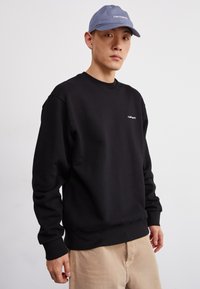 Black cotton sweatshirt with long sleeves and a crew neck, featuring a small white logo on the chest. Paired with light-colored trousers.