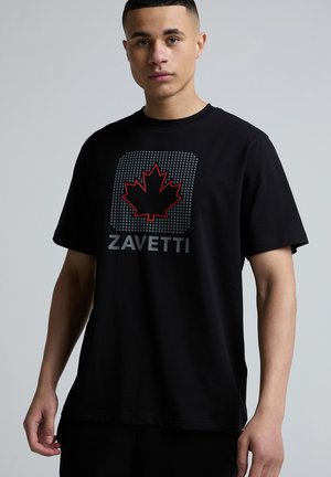 Nuori mies, joka pukeutunut mustaan T-paitaan, jossa on punainen vaahteranlehti ja harmaat "ZAVETTI"-tekstityypit pistekuvioisella neliötaustalla, neutraali harmaa tausta.