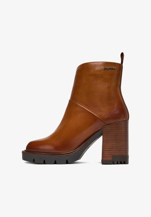 Stivaletto alla caviglia in pelle marrone con tacco in legno spesso, punta arrotondata e finitura liscia. Presenta un tirante laterale e un piccolo logo decorativo.