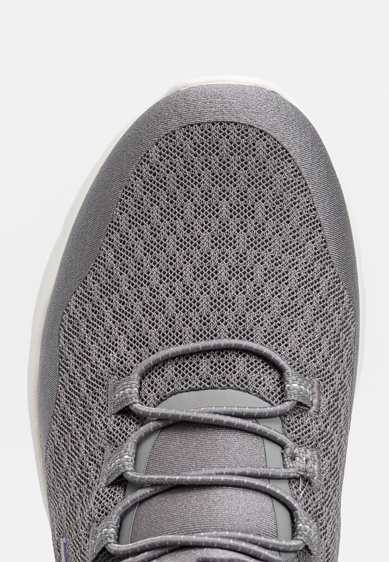 Zapatilla deportiva gris con parte superior de malla transpirable, cordones redondos y una puntera texturizada de material sintético. Media suela blanca y sutiles acentos morados.