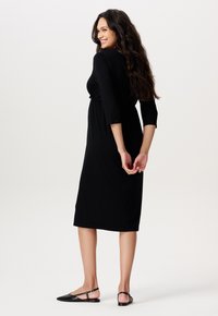 Schwarzes knielanges Kleid mit Dreiviertelärmeln, tailliertem Schnitt und glattem Stoff. Wird mit schwarzen flachen Schuhen getragen, Modell seitlich gezeigt.