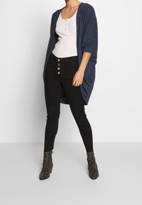 Frau in einem weißen ärmellosen Top, einer offenen cardigan in marineblau, schwarzen hochtaillierten Skinny Jeans mit Knöpfen und Schlangenleder-Stiefeletten.