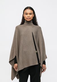 Γυναίκα με μακριά σγουρά μαλλιά, φορώντας ένα καφέ παλτό με ζιβάγκο πάνω από ένα μαύρο σύνολο, που στέκεται μπροστά από έναν απλό ανοιχτό φόντο.