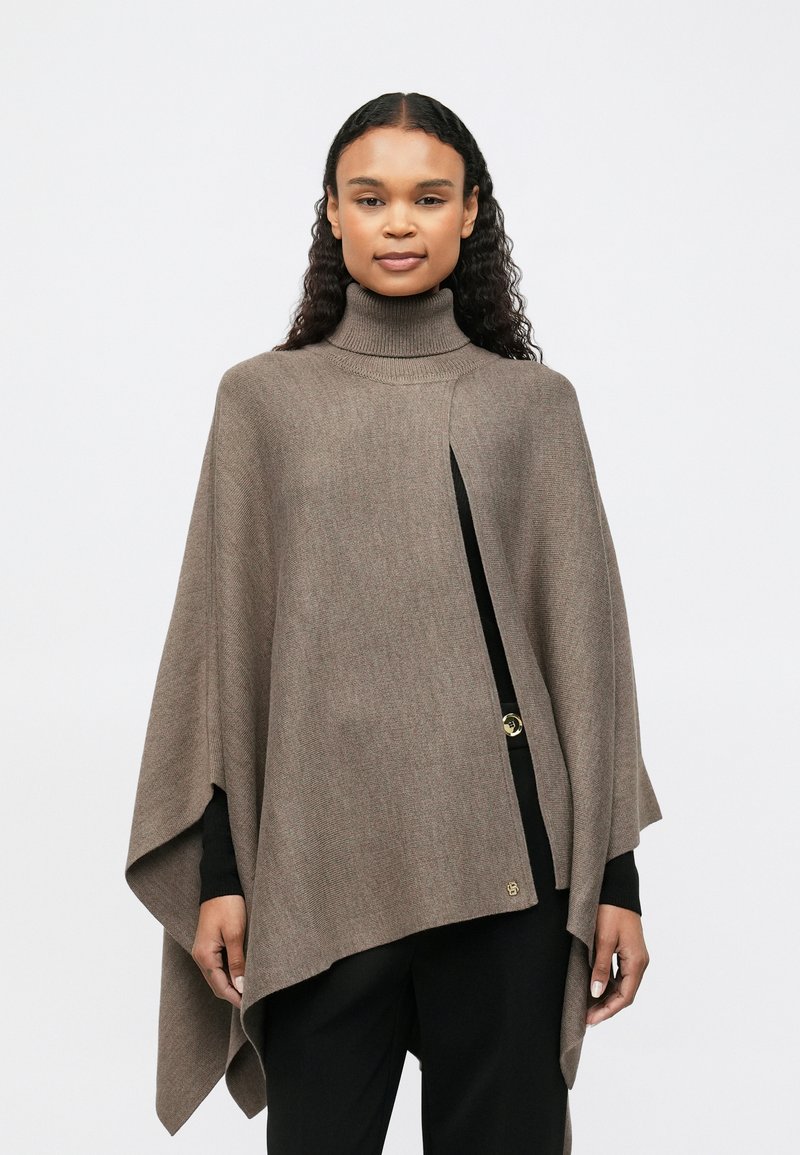 Γυναίκα με μακριά σγουρά μαλλιά, φορώντας ένα καφέ παλτό με ζιβάγκο πάνω από ένα μαύρο σύνολο, που στέκεται μπροστά από έναν απλό ανοιχτό φόντο.