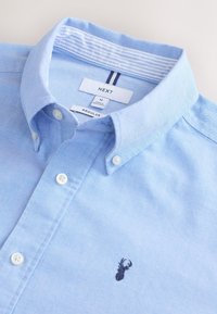 Lichtblauw overhemd met knoopsluiting in een getextureerde stof en een kraag, voorzien van een klein zwart geborduurd logo en witte knopen.