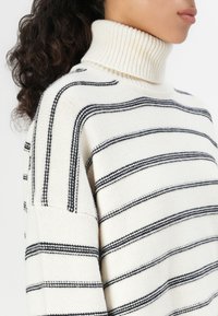 Gestreifter Rollkragenpullover in Creme und Marineblau, mit einem gerippten Kragen und weichem, strukturiertem Stoff, der horizontale Streifen über den Körper zieht.