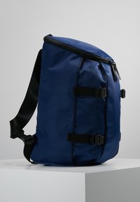 Niebieska torba na ramię z czarnym regulowanym paskiem, dwoma bocznymi zapięciami i zamkiem błyskawicznym, ustawiona pionowo na białym piedestale na szarym tle.