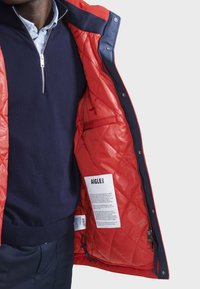 Rote Steppjacke mit dunkelblauer Fütterung, silbernem Reißverschluss und Pflegeetikett. Das Innere hat eine glatte Textur mit einem Rautenmuster und gesicherten Taschen.
