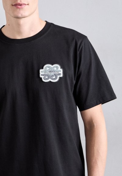 T-shirt en coton noir à manches courtes, avec un patch texturé blanc et gris portant l'inscription "Filling Pieces" sur le côté supérieur gauche de la poitrine.