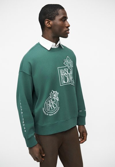 Νεαρός άνδρας που φοράει πράσινο φούτερ με λευκά γραφικά λογότυπα και κείμενο, φορεμένο πάνω από ένα λευκό πουκάμισο με γιακά, στέκεται μπροστά σε λευκό φόντο.