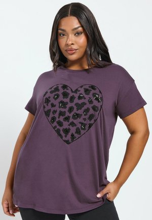 Mujer con cabello largo y oscuro, vistiendo una camiseta morada con un gran diseño de corazón de estampado de leopardo negro, de pie con una mano en la cadera.