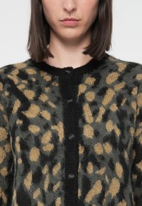 Torso de mulher em um cardigã felpudo e abotoado com um padrão abstrato de leopardo nas cores preto, cinza e bege, cortado abaixo do nariz, com cabelo castanho escuro liso.