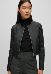 Svart läderbomberjacka med rund halsringning, dragkedjestängning och sidfickor, tillsammans med en svart polotröja och mönstrade grå byxor.