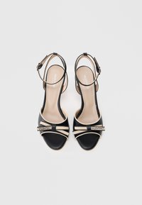 Czarne skórzane sandały mają otwarty nosek, pasek wokół kostki z klamrą oraz kontrastujące kremowe akcenty, w tym logo i paski na przodzie.