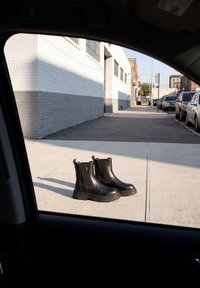 Een paar zwarte leren enkellaarzen op het zonovergoten stedelijke trottoir naast geparkeerde auto's, gezien door een autoruit.