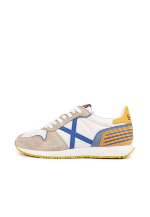 Zapatillas deportivas en blanco con ante beige y detalles en azul. Presentan una superficie texturizada, suela exterior amarilla y detalle de rayas en el talón.