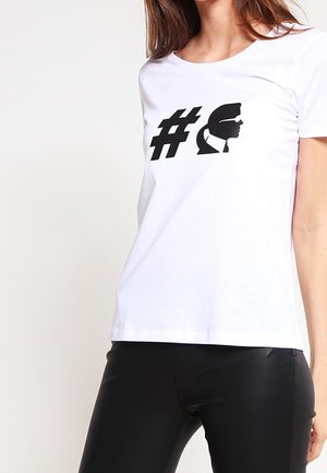 Nainen pukeutunut valkoiseen t-paitaan, jossa on musta hashtag-symboli ja miehen sivuprofiilin siluetti aurinkolaseissa ja solmiossa.