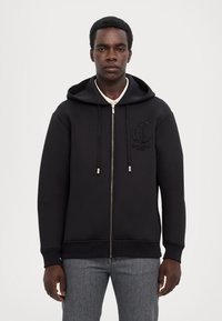 Felpa nera con zip e logo testurizzato sul lato sinistro del petto, cappuccio con cordini e polsini a costine. Tessuto liscio e vestibilità comoda.