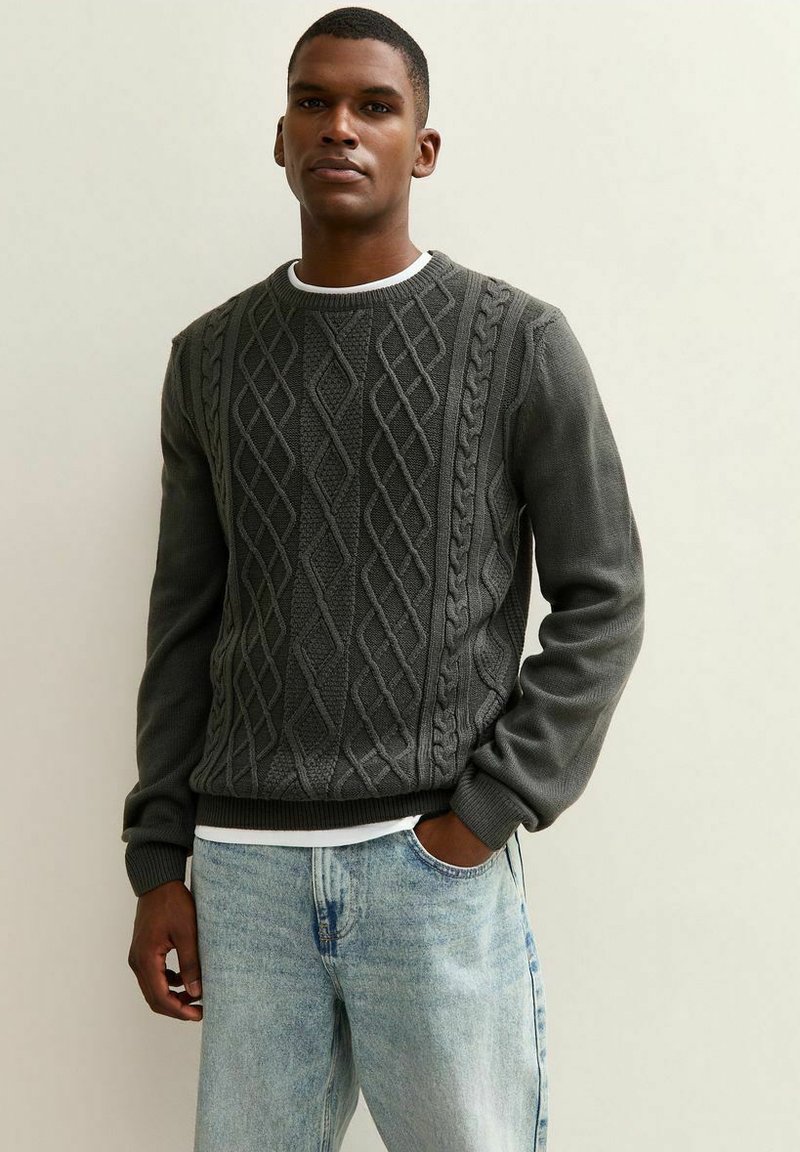 Suéter gris de punto con un patrón de diamante y trenzas texturizadas, con puños y dobladillo acanalados, llevado sobre una camisa blanca y combinado con pantalones de mezclilla claros.