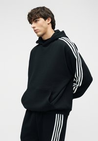 Giovane uomo con felpa nera con cappuccio e pantaloni coordinati con strisce bianche sulle maniche e sulle gambe dei pantaloni, in piedi con le mani dietro la schiena.