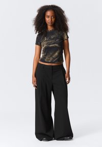 Kvinne med krøllete hår har på seg en svart og brun t-skjorte med dragegrafikk som er stukket inn i høytlivde, vide svarte bukser og svarte sko.