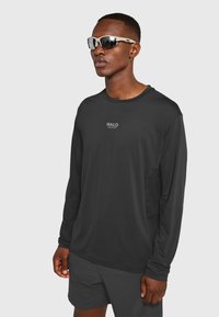 Czarna sportowa koszulka z długim rękawem z logo "HALO", z okrągłym dekoltem i elastycznego materiału, w zestawieniu z ciemnymi szortami i okularami przeciwsłonecznymi.