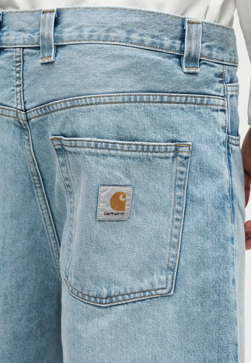Jean en denim bleu clair avec un patch logo Carhartt sur la poche arrière, porté par une personne en chemise blanche.