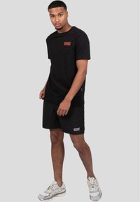 Sort bomulds t-shirt med rødt logo på brystet, parret med sorte shorts med hvid tekst "WRONG FRIENDS". Modellen har grå sneakers på.