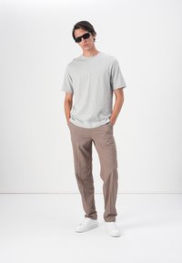 Un tricou gri din bumbac asociat cu pantaloni bej croiți și adidași albi. Modelul poartă ochelari de soare negri, stând pe un fundal alb.