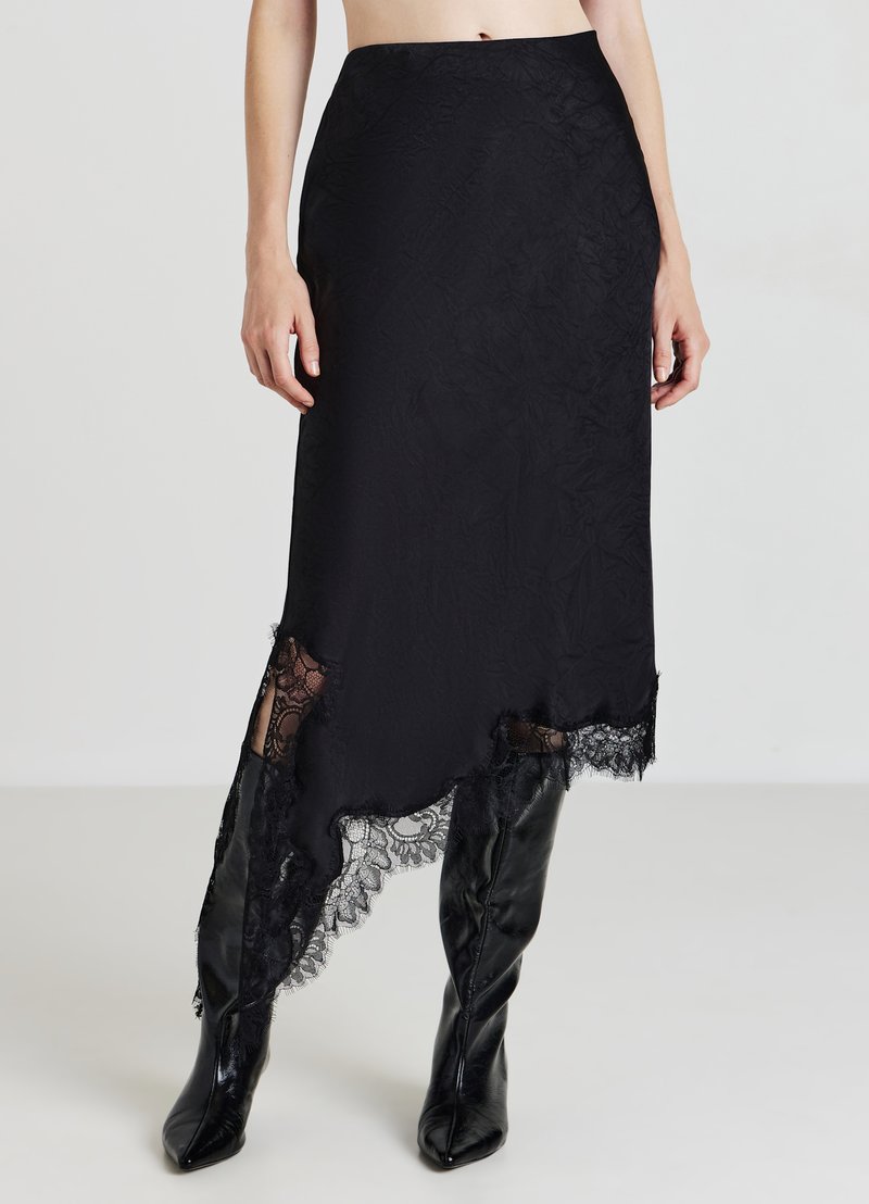 Jupe midi noire avec un ourlet asymétrique, ornée de dentelle au bas et d'une surface texturée. Portée avec des bottes noires montantes.