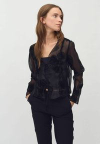Bluză neagră transparentă cu model floral, margini volănașe și detaliu cu siret. Mâneci lungi, material ușor, suprapusă peste un top închis la culoare.